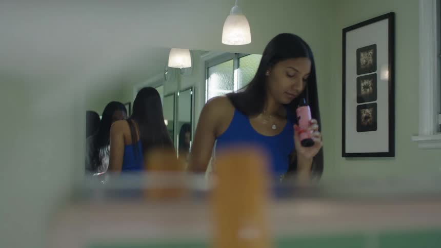 A young woman is standing in a room with her back to a mirror as another female hands her a bottle and she smells it and looks inside it