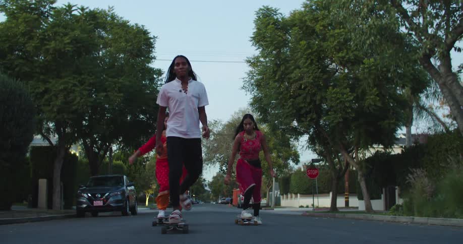 Man with long hair white t shirt black pants and two women with red sari skateboard in city street