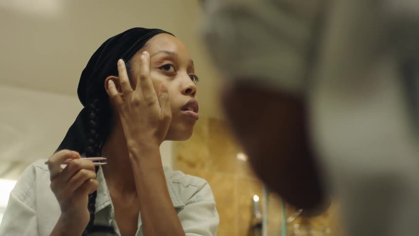 Woman uses a pincer to trim her eyebrows in front of a mirror inside a bathroom