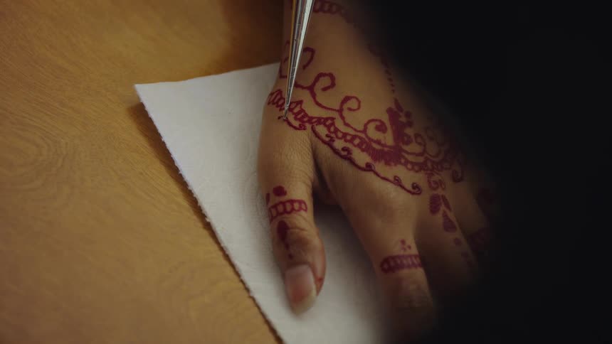 Artists drawing red henna ink tattoo on the hand of a woman over a wooden table