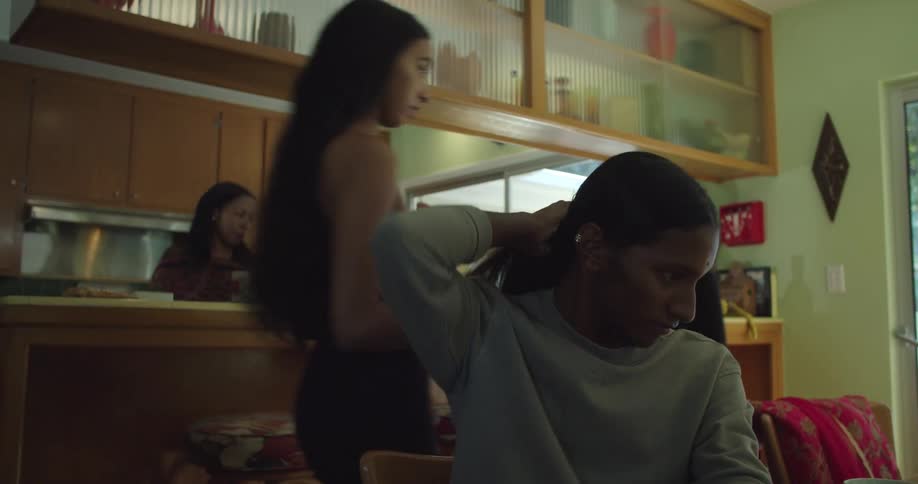 A teenager sits at a table in a restaurant as people walk past her carrying dishes and two women cook in the kitchen