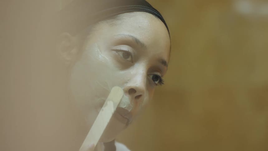 Young woman applying cream with a spatula on her face in front of a mirror at home