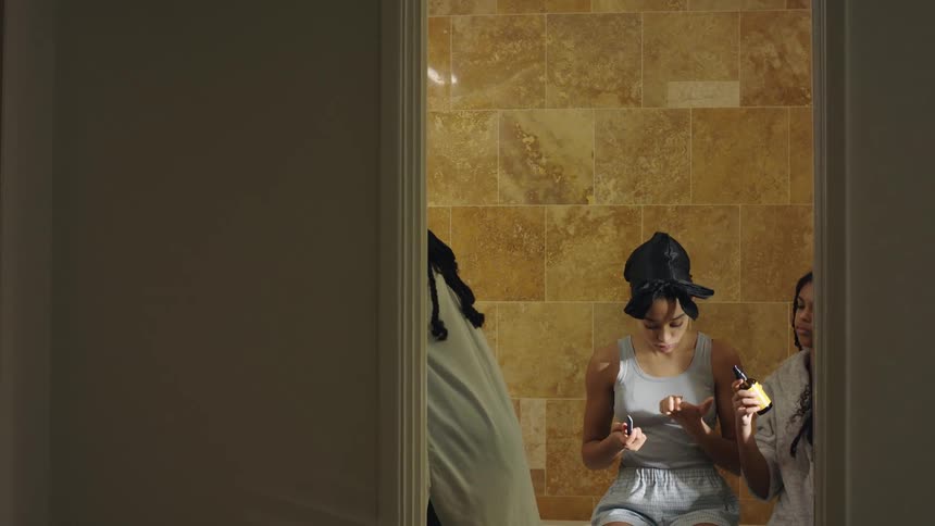 A girl sitting on a bathtub files her nails while the girl next to her puts serum in her hair and another girl stands in front of them