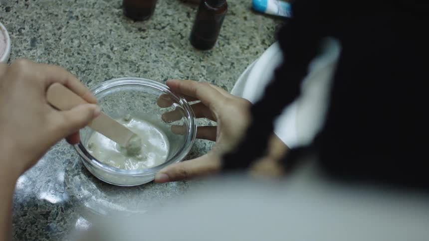 Woman stirs clay mixture in small bowl near bathroom sink