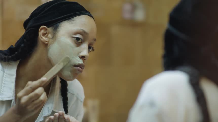 A black woman uses a tongue depressor to spread a face mask on her cheek