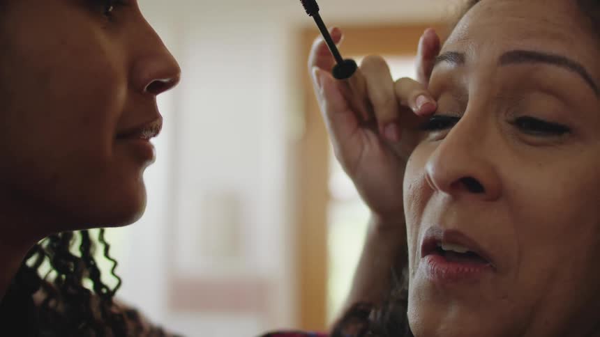 Young woman applies makeup on the eyelashes of a startled woman at home