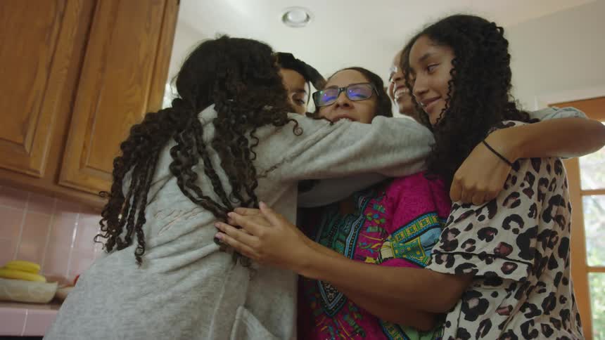 Four teenage girls are group hugging an older woman as they stand in a kitchen