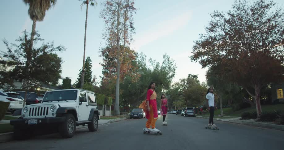 Three people skateboard down the neighborhood street with the girls in traditional sari