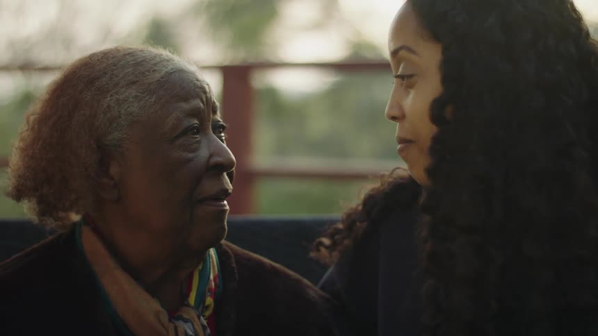 Young woman with long curly hair talks to elderly woman with gray hair and green yellow and red scarf touching her nose