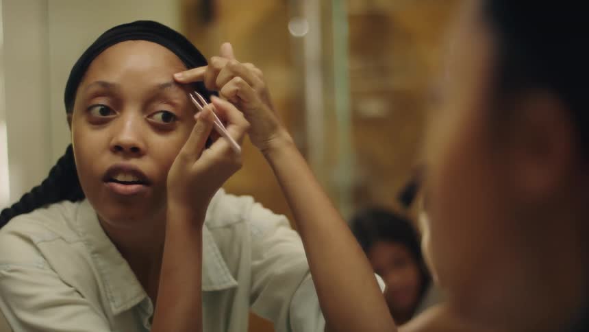 A woman tweezes her eyebrow while talking to her sisters in the mirror reflection