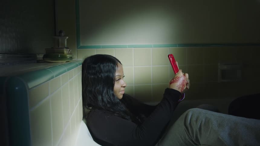 A teenage girl sits in a bathtub and takes selfies on her cell phone