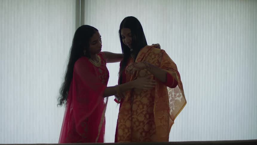Two women are standing indoors and wearing saris