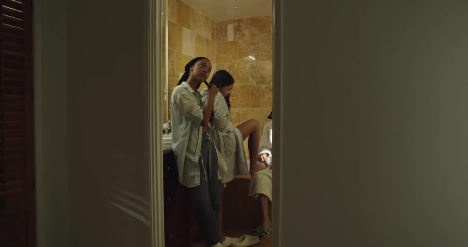 Three young girls wearing white bathrobes in the bathroom doing their nails and braiding hair