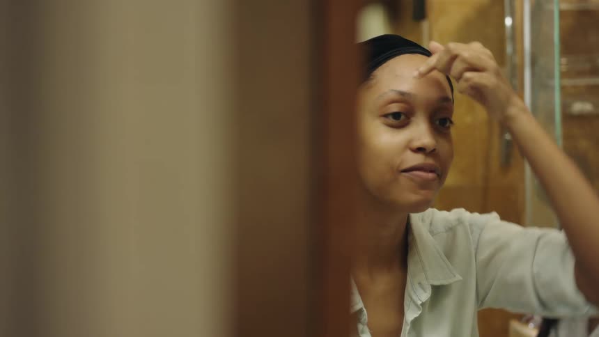 Woman uses a pincer to trim her eyebrows in front of a mirror inside a bathroom with her friends