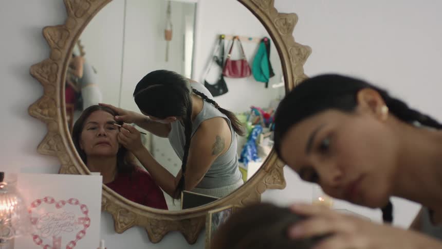 Woman with braids gray under shirt puts makeup on elderly woman wearing red sweatshirt looking in mirror in beauty salon