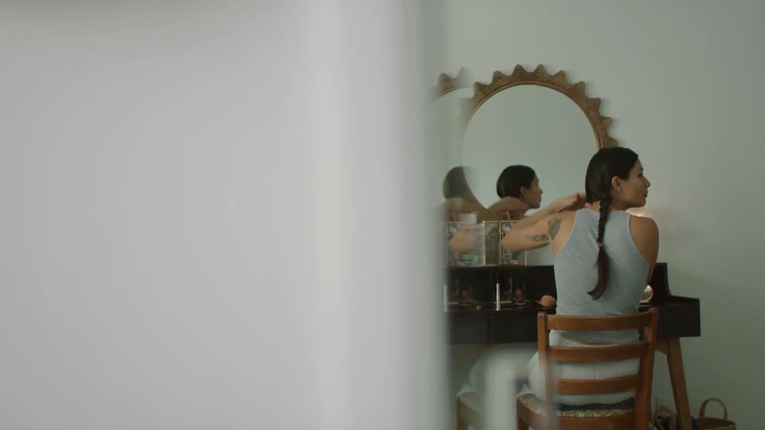 Woman sits at vanity in front of mirror braiding her hair