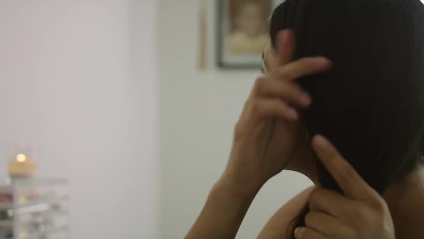 Woman with black wavy hair and gray undershirt does her hair looking in mirror