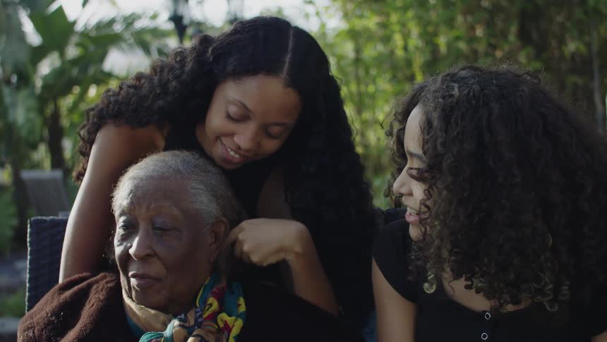 Two young black women are outside with their grandmother showing her affection