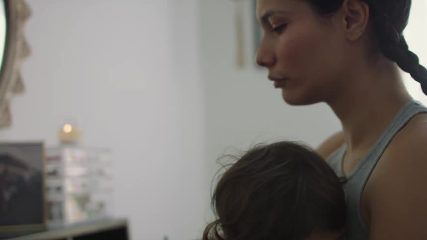 Woman with braids and gray undershirt breastfeeds toddler girl while putting on make up looking in mirror