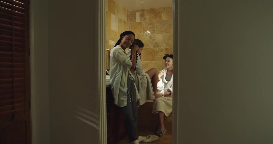 Three young girls wearing white bathrobes in the bathroom doing their nails and braiding hair