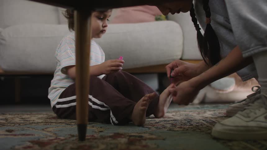 A mom squatting down painting her toddlers toes while she sits on the carpet