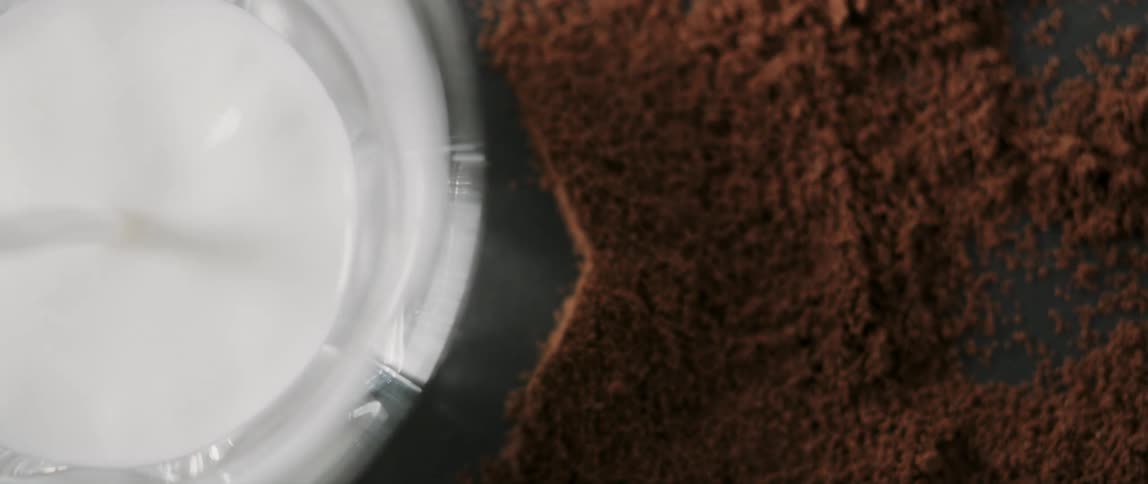 Top-down view of a glass being filled with milk next to loose fresh coffee grounds