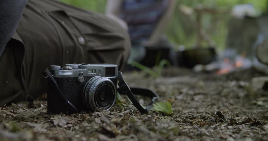 A camera sits on the ground while in the background a woman tends to something near a campfire