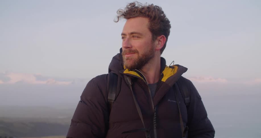 Man with beard black coat and backpack looks over his shoulder smiling on the shore at dusk