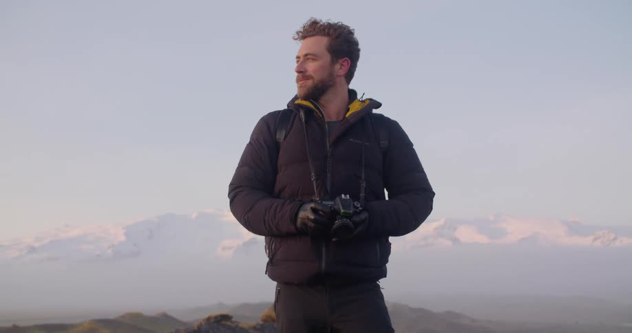 A man holding a camera and looking around in awe stands on a high ridge with mountains behind him