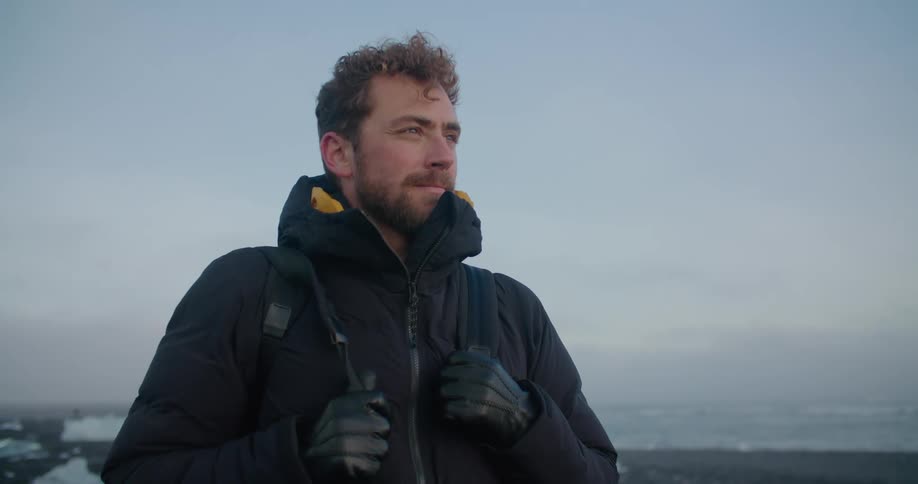 A photographer in a winter coat looks out into the distance while standing on a beach covered in blocks of ice in the evening