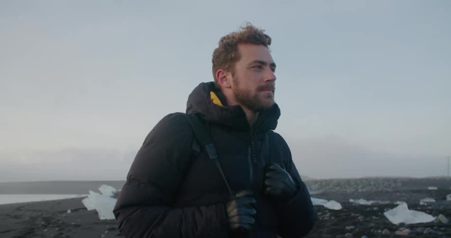 Man with beard black coat and gloves walking on the shore looking around at dusk