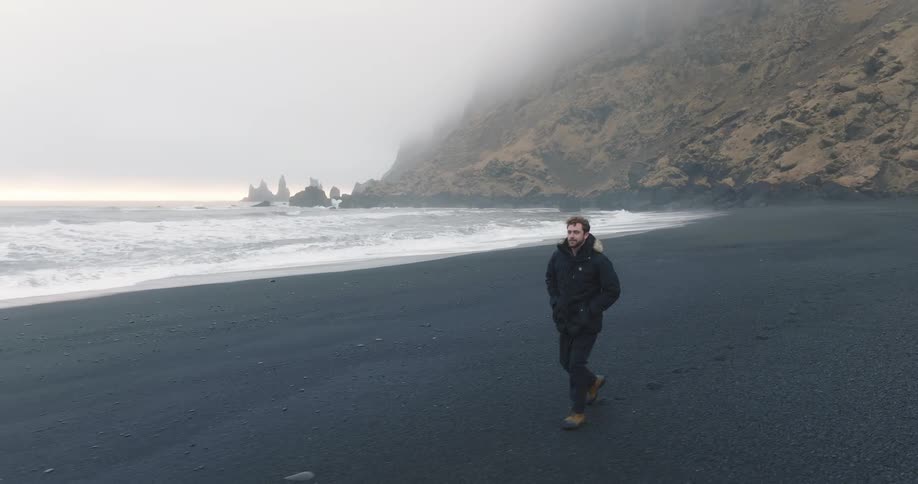 Young man walking on the black sand beach next to the ocean close to the mist covered mountains