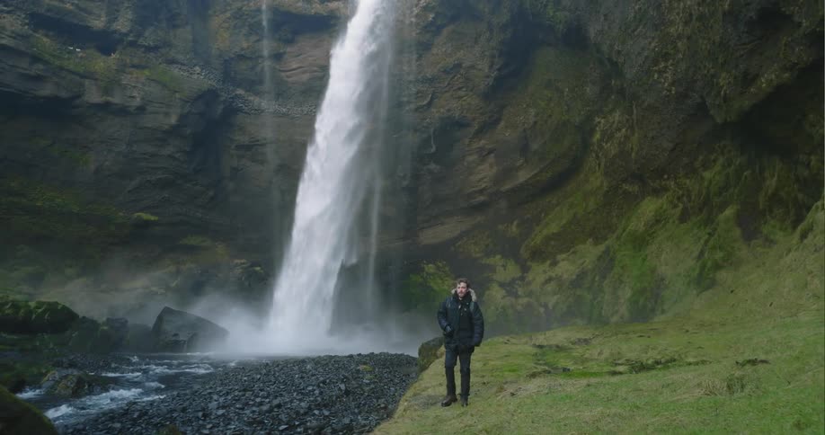 A man wearing a coat is walking near a waterfall flowing down the side of a cliff