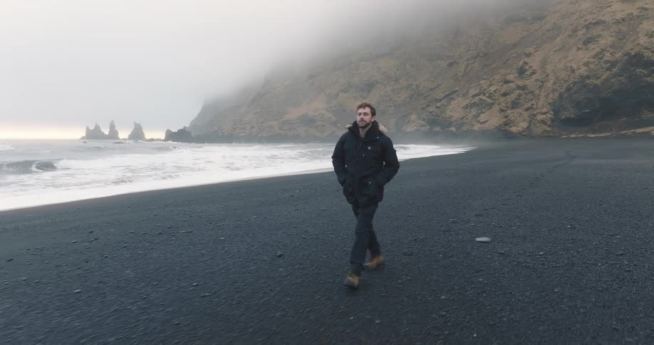 Young man walking on the black sand beach next to the ocean close to the mist covered mountains