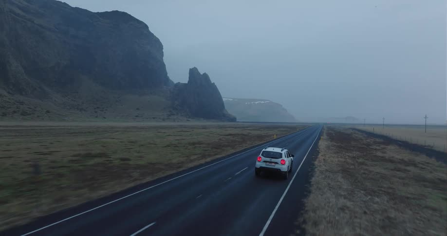 White car drives on highway with high cliff and fog in the background