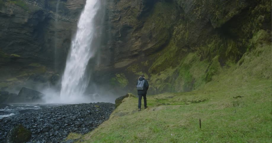 A man walks through the wilderness towards a waterfall before stopping to observe it