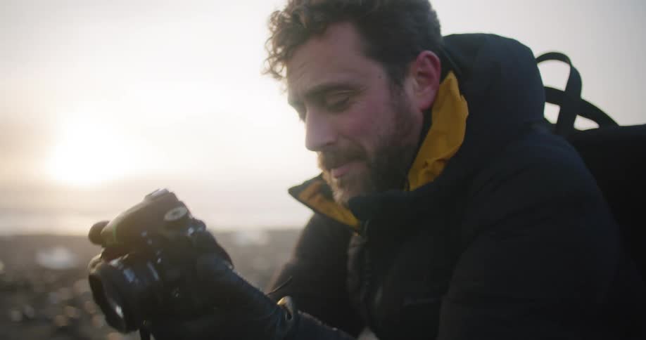 Young bearded man takes photos with a camera on the coast at sunset