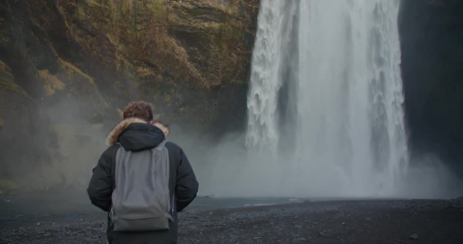 Man with black coat gray backpack walks to waterfall cascading down cliffside