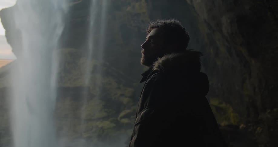 A man in a heavy coat standing next to a waterfall in nature
