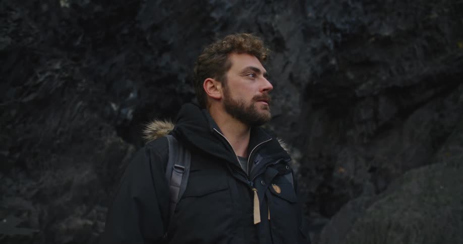 A man with a backpack on stands in a mountain range looks out at the landscape