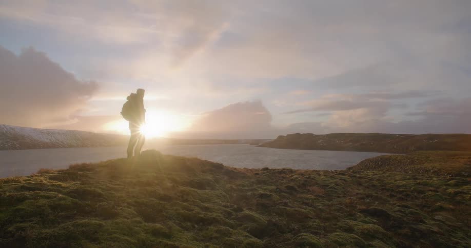 A man is standing at the side of a lake as the sun is shining brightly on the horizon