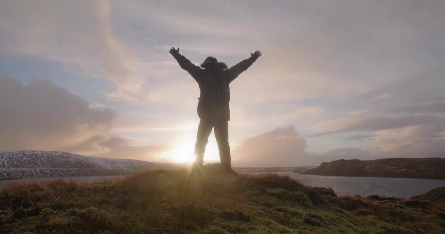 A man standing on top of a hill by a lake raises his hands in the air and cheers