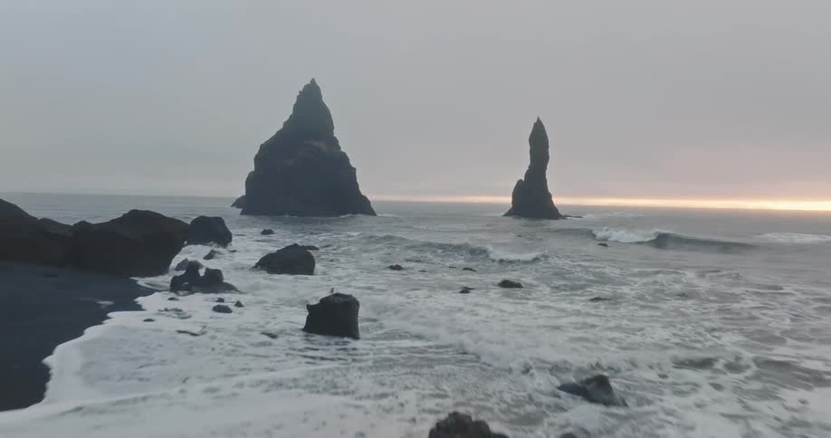 Rock formations jut out the ocean near a beach with dark soil on a cloudy day