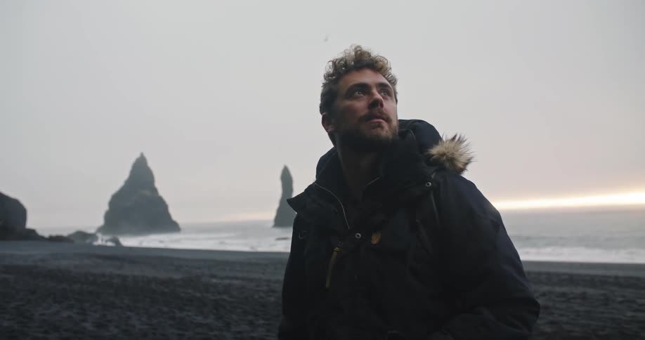 A man stands on a rocky beach looking away from the water