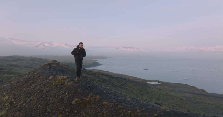 A man wearing a backpack is walking on top of a hill along the coast with mountains in the distance behind him