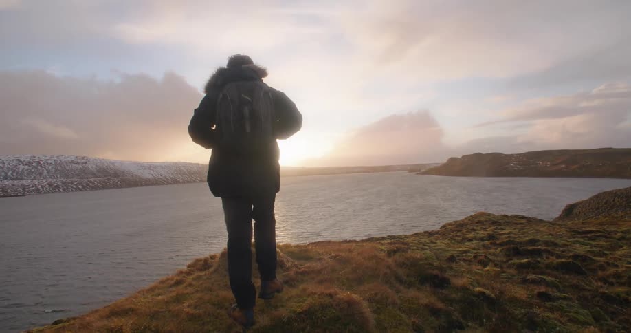 Young bearded man walks towards the coast close to an icelandic fjord at sunset