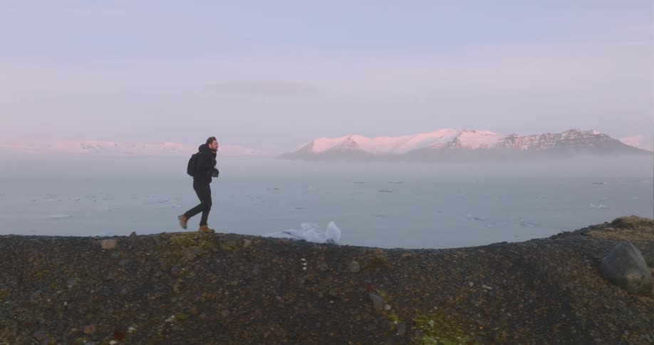 A man hikes along a hill on the shore with large icebergs
