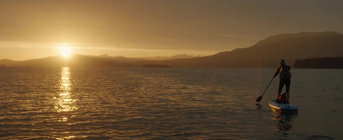 A person standing over a board paddling with an oar in the ocean close to the mountains at sunset