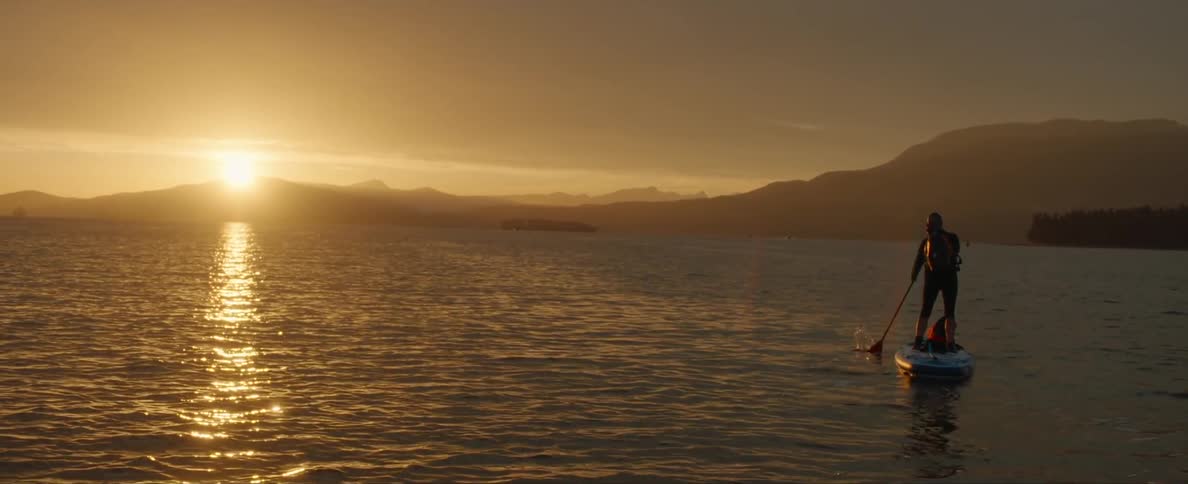 A person standing over a board paddling with an oar in the ocean close to the mountains at sunset