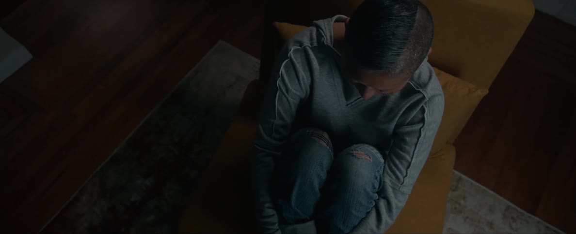 Sad young person sitting on a chair with arms around the legs inside a dimly lit room